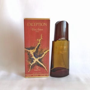 Puede incluir: Un conjunto de perfume con frasco y caja. La caja es roja con detalles dorados y la palabra "EXCEPTION" en la parte superior. El frasco es marrón con una tapa marrón oscuro. El texto "EXCEPTION Remy Latour Paris" es visible.