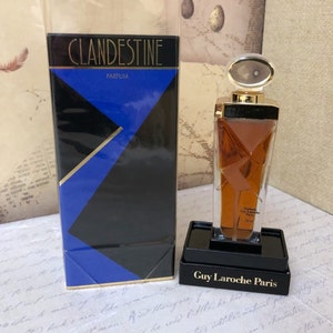Puede incluir: Una caja dorada y negra con el texto "Clandestine Parfum" en la parte delantera. Una botella de perfume de vidrio transparente con una tapa dorada y una etiqueta dorada que dice "Parfum Guy Laroche Paris 15 ml" está sentada sobre una base negra.