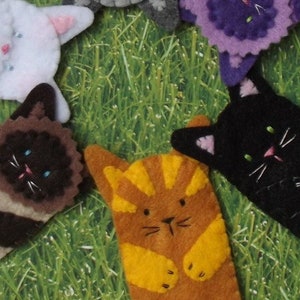 May include: Six felt finger puppets shaped like cats in different colors and patterns. The puppets are arranged in a circle on a green grassy background.
