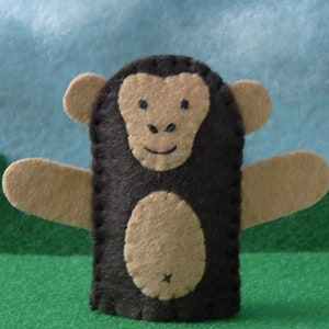 May include: A dark brown and tan felt finger puppet shaped like a monkey. The puppet features a rounded face with small ears, a central oval belly patch, and two outstretched arms. The facial features are stitched in black thread on the tan felt.