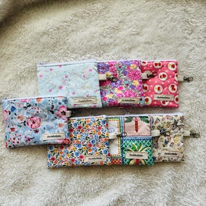 May include: Eight small zippered pouches with various floral and patterned fabric designs. The pouches are lined up in a row on a white fluffy surface. Each pouch has a small label that says "Charlotte Stitches".