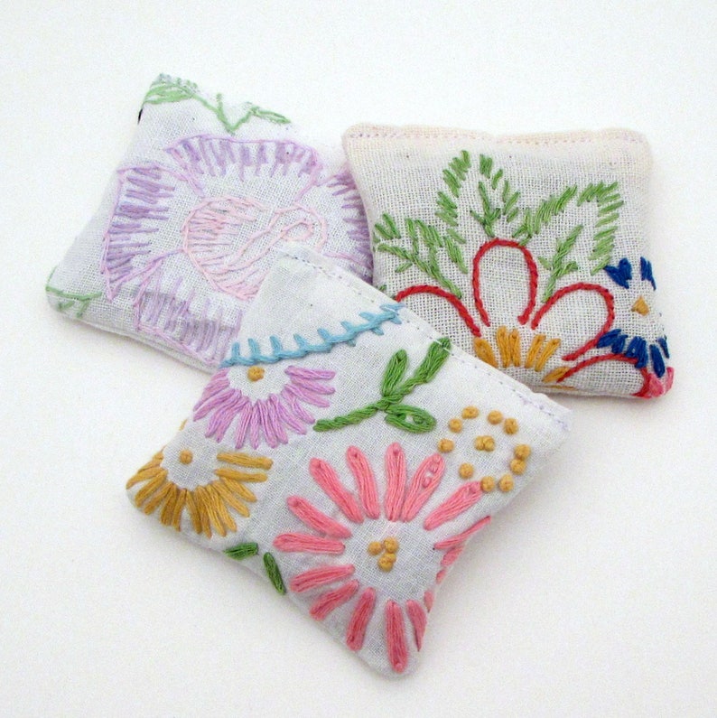 May include: Three white, square sachets with colorful floral embroidery. The embroidery features pink, yellow, green, and blue flowers. The sachets are arranged on a white background, showcasing the handmade details.