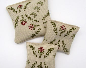 May include: Three beige cross-stitched sachets with a floral pattern in red and green. The sachets are square and appear to be filled with a fragrant material. The cross-stitch design features stylized flowers and leaves.