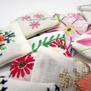 May include: Close-up of several small, white, embroidered fabric pouches. Each pouch features a different floral design in various colors, including pink, red, yellow, and green. The embroidery is detailed and appears handmade, showcasing a variety of stitches.