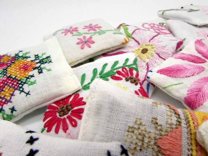 May include: Close-up of several small, white, embroidered fabric pouches. Each pouch features a different floral design in various colors, including pink, red, yellow, and green. The embroidery is detailed and appears handmade, showcasing a variety of stitches.