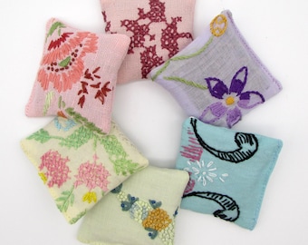 May include: A collection of six small, square, embroidered pillows in various pastel colors. Each pillow features a unique floral or abstract design in shades of pink, purple, blue, green, and yellow. The pillows are arranged in a circular pattern.