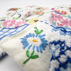 May include: Close-up of several small, white fabric sachets with colorful floral embroidery. The embroidery features pink, blue, yellow, and green thread, depicting various flower designs. The sachets are likely filled with potpourri or herbs.