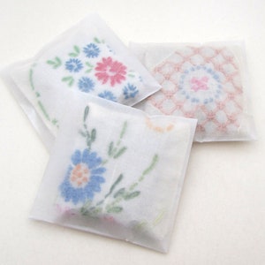 May include: Three white, square sachets with floral embroidery. One sachet has blue and pink flowers, another has a pink and blue geometric design, and the third has blue flowers with orange centers. The sachets are wrapped in translucent paper.