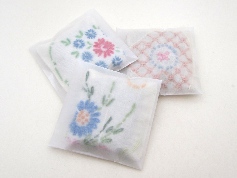 May include: Three white, square sachets with floral embroidery. One sachet has blue and pink flowers, another has a pink and blue geometric design, and the third has blue flowers with orange centers. The sachets are wrapped in translucent paper.