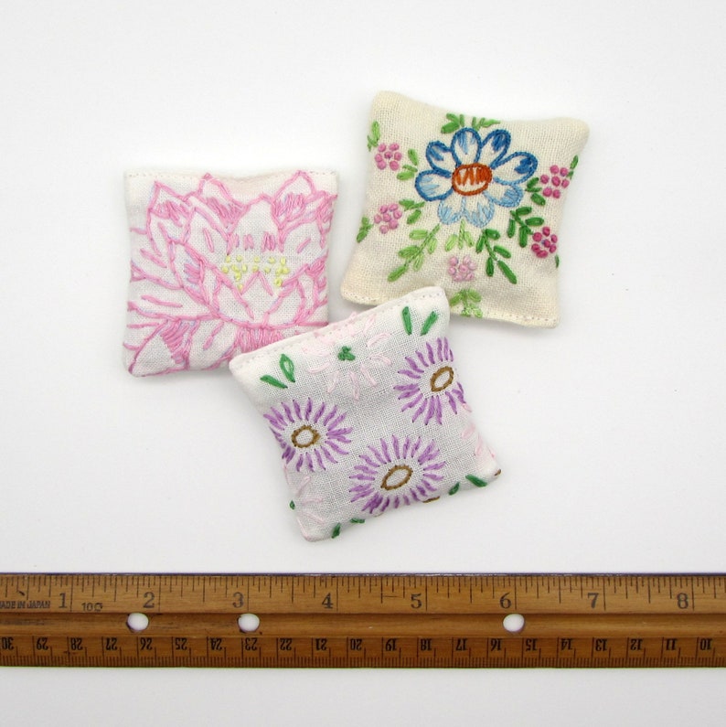 May include: Three small, square, embroidered pillows with floral designs. One pillow features a pink lotus flower, another has a blue flower with green leaves, and the third has purple daisies. The pillows are on a white background.