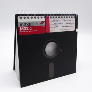 May include: A black floppy disk calendar with a spiral binding. The front features a red and white label with "maxell MD2-D" printed on it. A handwritten note on a red sticky note reads "Personal Consultant Migration Utilities (for IBM PC)."