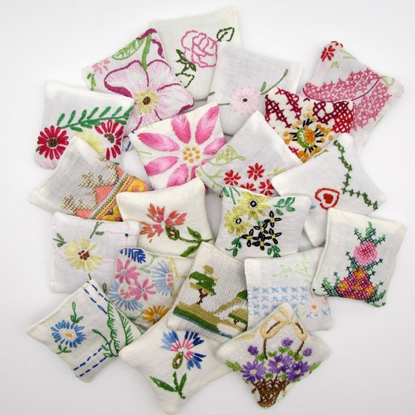 May include: A collection of small, square, white fabric pillows, each adorned with colorful, hand-embroidered floral designs. The embroidery features various flowers, leaves, and stems in shades of pink, red, yellow, and blue.