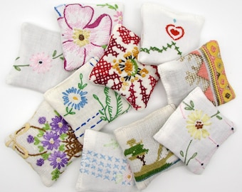 May include: A collection of small, white fabric sachets, each embroidered with a unique floral or geometric design. The embroidery features various colors, including pink, blue, yellow, and red, creating a handcrafted aesthetic. The sachets are arranged on a white surface.