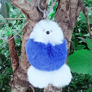 Könnte beinhalten: Ein weißer, flauschiger Schlüsselanhänger mit einem blauen Fellbommel. Der Schlüsselanhänger hat einen silbernen Ring und eine weiße Schleife.