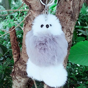 Könnte beinhalten: Ein grauer und weißer flauschiger Pompon-Schlüsselanhänger mit schwarzen Augen. Der Schlüsselanhänger ist an einer silbernen Kette befestigt.