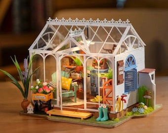 May include: A miniature greenhouse model with a white frame and glass panels. Inside, there are tiny plants, furniture, and decorative items. A small cart with flowers and the word "Welcome" is in front. The model includes a small blue door and window.