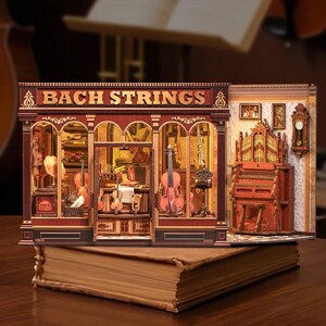 Può includere: Un modello in legno dettagliato di un negozio di musica, "Bach Strings", con facciata rossa e oro. Il negozio presenta violini in miniatura, un violoncello e un organo. Il modello è posto sopra un grande libro aperto.
