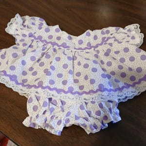 May include: A white and purple polka dot baby outfit with a ruffled top and matching bloomers. The outfit is trimmed with white lace and purple ribbon. The fabric is a lightweight cotton.