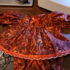 May include: A festive doll outfit featuring a burgundy and red holly leaf pattern. The top has puffed sleeves and a small orange bow. The outfit includes matching ruffled bloomers, trimmed with orange and silver lace.