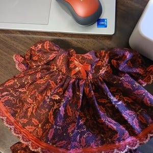 May include: A doll dress in shades of burgundy and red, featuring a pattern of leaves and gold accents. The dress has a ruffled hem and sleeves, with a small bow at the neckline. The dress is designed for a doll.