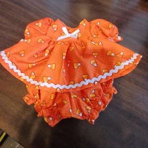 Peut inclure: Ensemble orange pour bébé avec un motif de bonbons. L'ensemble comprend un haut avec un nœud blanc et des manches à volants, ainsi qu'un short à volants assorti. Une bordure blanche festonnée orne les bords du haut et du short.