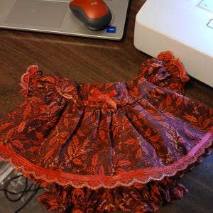 May include: A doll's dress and bloomers set. The set features a dark red fabric with a holly leaf pattern and red lace trim. A small red bow adorns the dress. The bloomers have a ruffled hem.