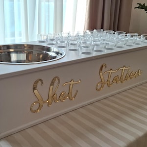 Può includere: Un'elegante stazione per shot in legno bianco con un bacino per il ghiaccio in acciaio inossidabile e numerosi bicchierini. Le parole "Shot Station" sono visualizzate in lettere dorate sul davanti. La stazione è su una tovaglia a righe.