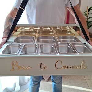 Könnte beinhalten: Ein hellgraues Cannoli-Serviertablett mit Edelstahlbehältern. Das Tablett hat goldfarbene Schriftzüge mit der Aufschrift "Born to Cannoli". Es wird von einer Person mit einem schwarzen Gurt gehalten.
