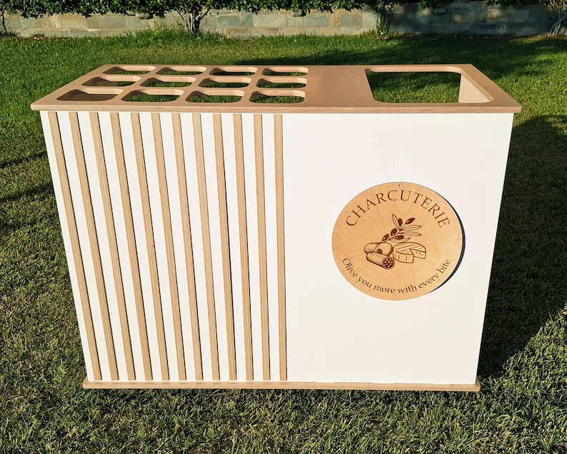 Puede incluir: Una estaci&oacute;n de servicio de charcuter&iacute;a blanca y de madera con un dise&ntilde;o de listones en un lado. La estaci&oacute;n presenta un letrero circular de madera que dice "Charcuter&iacute;a" con el texto "Olive you more with every bite."
