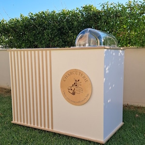 Puede incluir: Un expositor de charcuter&iacute;a blanco y de madera con una cubierta de c&uacute;pula transparente. El expositor presenta un letrero redondo de madera con la palabra "CHARCUTERIE" y un dise&ntilde;o de rama de olivo. El expositor est&aacute; sobre un c&eacute;sped verde.