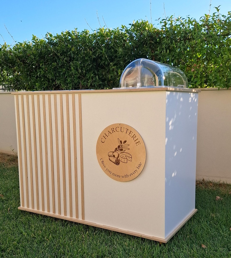 Puede incluir: Un expositor de charcuter&iacute;a blanco y de madera con una cubierta de c&uacute;pula transparente. El expositor presenta un letrero redondo de madera con la palabra "CHARCUTERIE" y un dise&ntilde;o de rama de olivo. El expositor est&aacute; sobre un c&eacute;sped verde.