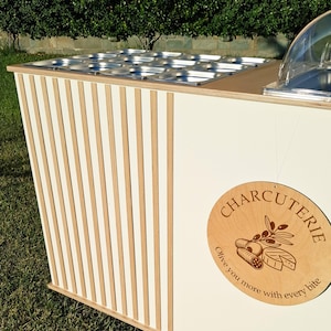 Puede incluir: Un carrito de charcuter&iacute;a blanco y de madera con un dise&ntilde;o a rayas. El carrito presenta un letrero de madera que dice "Charcuter&iacute;a" con ilustraciones de alimentos. Se ven bandejas de alimentos de acero inoxidable.