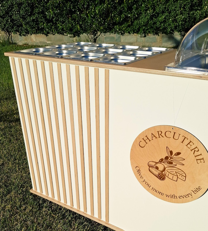 Puede incluir: Un carrito de charcuter&iacute;a blanco y de madera con un dise&ntilde;o a rayas. El carrito presenta un letrero de madera que dice "Charcuter&iacute;a" con ilustraciones de alimentos. Se ven bandejas de alimentos de acero inoxidable.