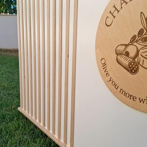 Puede incluir: Una estructura de madera con listones verticales de madera clara que se alternan con paneles blancos. Un letrero circular de madera con la palabra "CHARC" y un dise&ntilde;o de rama de olivo es visible. El letrero tambi&eacute;n incluye el texto "Olive you more wi..."