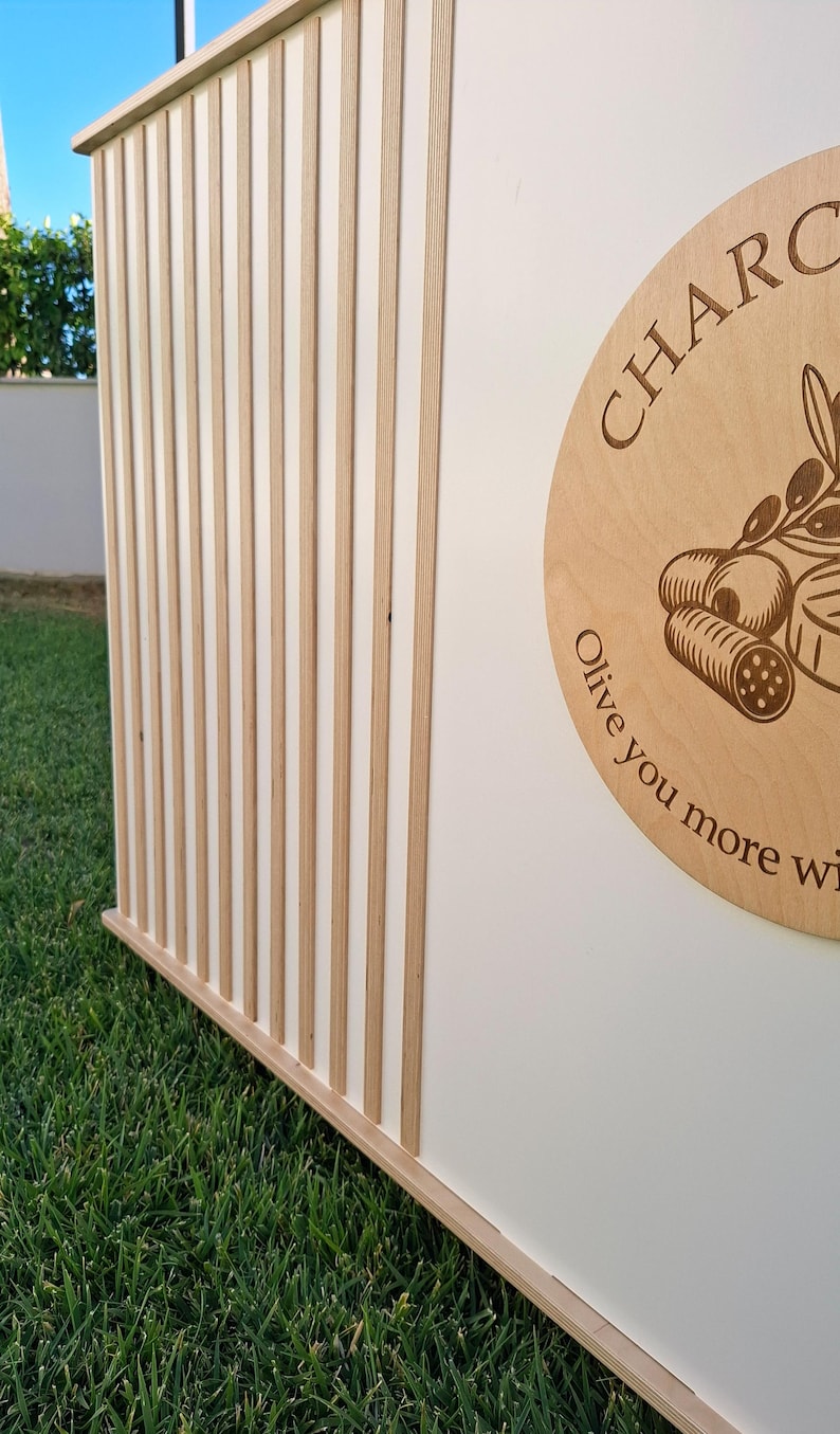 Puede incluir: Una estructura de madera con listones verticales de madera clara que se alternan con paneles blancos. Un letrero circular de madera con la palabra "CHARC" y un dise&ntilde;o de rama de olivo es visible. El letrero tambi&eacute;n incluye el texto "Olive you more wi..."