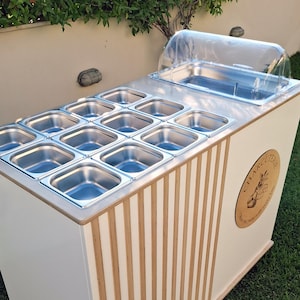 Puede incluir: Un carrito de charcuter&iacute;a blanco y madera, con una parte superior de acero inoxidable y m&uacute;ltiples recipientes para alimentos. Tiene una cubierta de pl&aacute;stico transparente y un emblema de madera con el texto "CHARCUTERIE". El carrito est&aacute; al aire libre sobre c&eacute;sped.