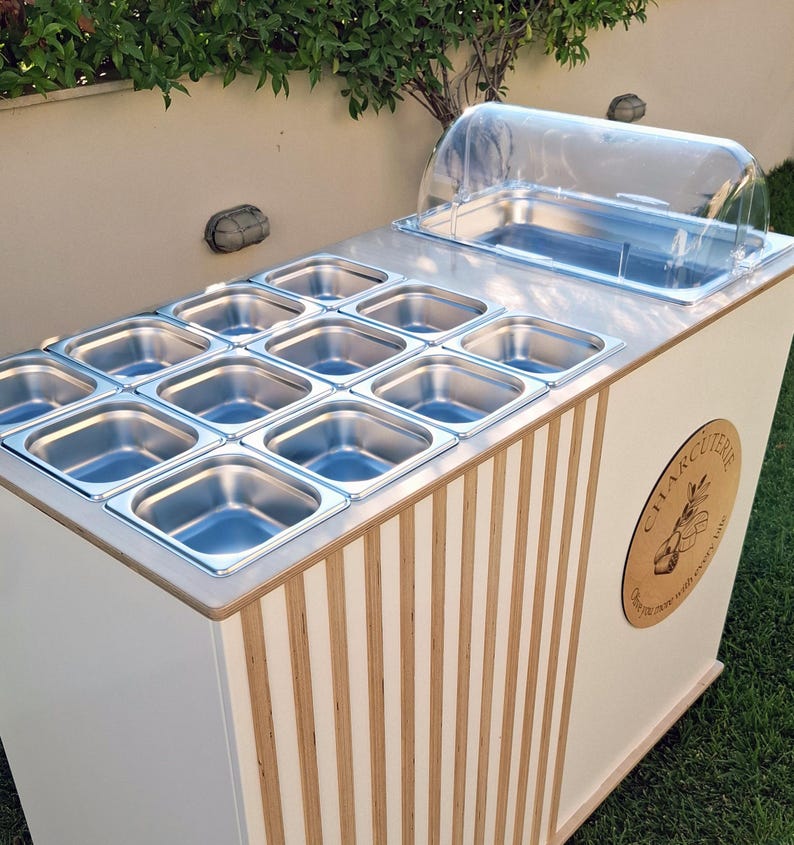 Puede incluir: Un carrito de charcuter&iacute;a blanco y madera, con una parte superior de acero inoxidable y m&uacute;ltiples recipientes para alimentos. Tiene una cubierta de pl&aacute;stico transparente y un emblema de madera con el texto "CHARCUTERIE". El carrito est&aacute; al aire libre sobre c&eacute;sped.