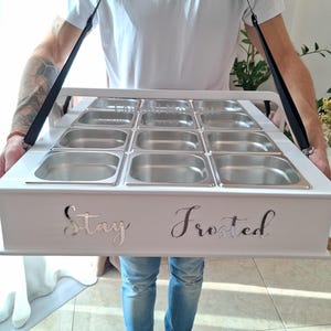 Op de afbeelding: Een wit houten serveerblad met een zwarte draagriem, voorzien van negen vierkante roestvrijstalen inzetstukken in een raster. De voorzijde van het blad toont de tekst "Stay Frosted" in een zilverkleurig, cursief en reflecterend lettertype.