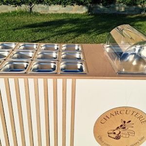 Puede incluir: Un carrito de servicio de charcuter&iacute;a blanco y de madera, con recipientes de alimentos de acero inoxidable y una c&uacute;pula de pl&aacute;stico transparente. El frente del carrito est&aacute; decorado con listones de madera verticales y un letrero redondo de "Charcuter&iacute;a".