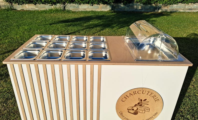 Puede incluir: Un carrito de servicio de charcuter&iacute;a blanco y de madera, con recipientes de alimentos de acero inoxidable y una c&uacute;pula de pl&aacute;stico transparente. El frente del carrito est&aacute; decorado con listones de madera verticales y un letrero redondo de "Charcuter&iacute;a".