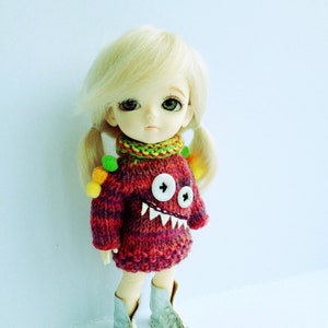 May include: A yellow doll with blonde hair wearing a red, green, and yellow knitted sweater with a white monster face on the front. The doll is wearing silver boots.