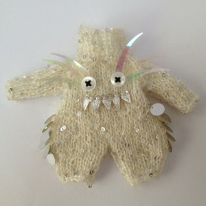 May include: A white knitted onesie with silver sequins and iridescent feather accents. The onesie has two black buttons for eyes and a silver jagged tooth design for a mouth.