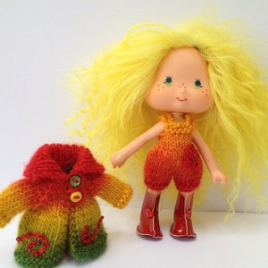 May include: A yellow haired doll wearing a red, orange, and yellow knitted jumpsuit with red boots. The doll is holding a red, orange, and green knitted jacket with a button closure.