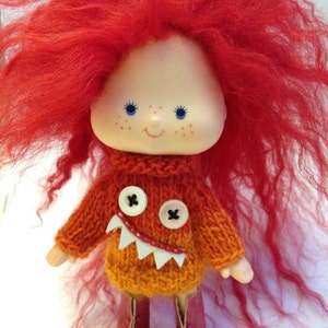 May include: A red-haired doll with blue eyes and a knitted orange sweater with a monster face. The sweater has white teeth and two black buttons for eyes.
