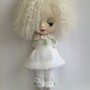 Puede incluir: Una muñeca blanca con el pelo blanco rizado que lleva un vestido y pantalones de punto blanco con detalles florales. La muñeca tiene un ojo azul y un ojo negro.