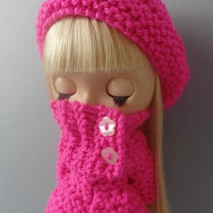 May include: A pink knitted doll outfit with a matching beret and two white buttons. The doll has blonde hair and closed eyes.