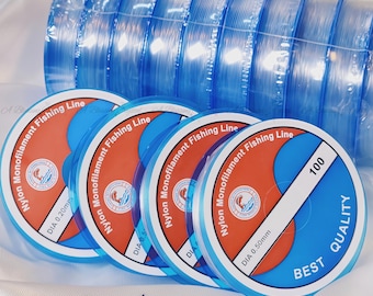 Può includere: Primo piano di una bobina di lenza da pesca in nylon monofilamento trasparente. La bobina è etichettata "Nylon Monofilament Fishing Line" e ha un diametro di 0,50 mm. La bobina è anche etichettata "Best Quality 100".