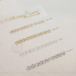 Könnte beinhalten: Nahaufnahme von Sprungringen in vier verschiedenen Ausführungen: 14 Karat Gold, 18 Karat Gold, Silber und Rhodium. Die Sprungringe sind in Reihen auf einer weißen Oberfläche angeordnet.