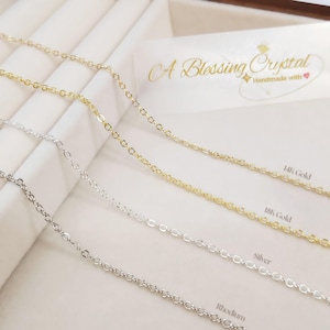 Peut inclure: Gros plan sur quatre fines chaînes de collier en or, argent et rhodium. Les chaînes sont présentées sur une surface blanche, avec le texte "A Blessing Crystal" et "Handmade with" visible en arrière-plan. Les chaînes sont étiquetées "14K Gold", "18K Gold", "Silver" et "Rhodium".
