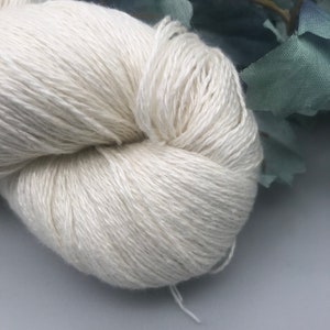 May include: A close-up of a skein of off-white yarn. The yarn is tightly wound and has a soft, textured appearance. The background includes a few blue-green fabric leaves.